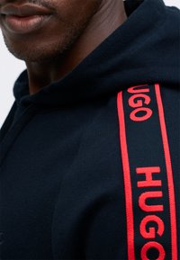 Detailný záber osoby v tmavom mikinovom hrube s červeným prúžkom s logom "HUGO" na ramene.