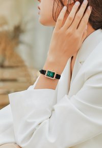 Reloj de pulsera de cuero negro con una carátula rectangular que presenta una esfera verde y acentos en oro rosa, engastada con pequeñas piedras transparentes alrededor del bisel.