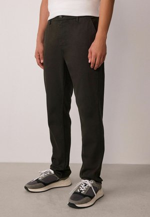 Dunkelgrüne Baumwollhose mit schmalem Schnitt, mit einem Frontknopfverschluss, Seitentaschen und einer glatten Textur. Kombiniert mit grauen Sneakers.