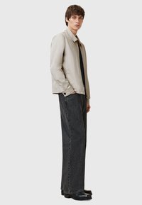 Beige bomberjack met een gladde textuur, staande kraag en drukknopen aan de voorkant, gedragen met losse donkere jeans en zwarte schoenen.