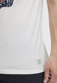 Kortermet hvit T-skjorte laget av myk bomull. Har et grafisk trykk i fargene oransje, blå og hvit. Inkluderer en logomerking ved kanten.