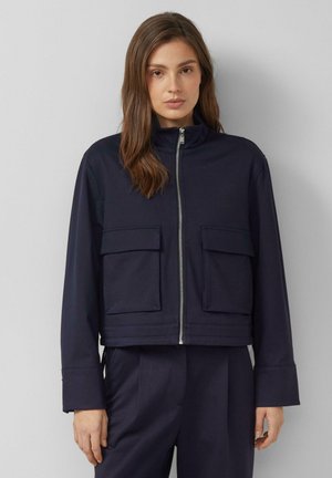 Femme aux cheveux bruns mi-longs portant une veste zippée bleu marine avec de grandes poches avant et un pantalon assorti bleu marine.