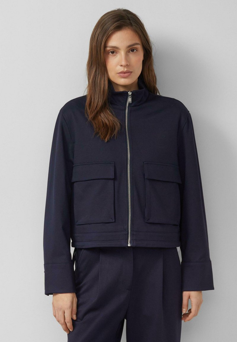 Frau mit schulterlangem braunem Haar, die eine marineblaue Jacke mit Reißverschluss und großen Vordertaschen sowie eine passende marineblaue Hose trägt.