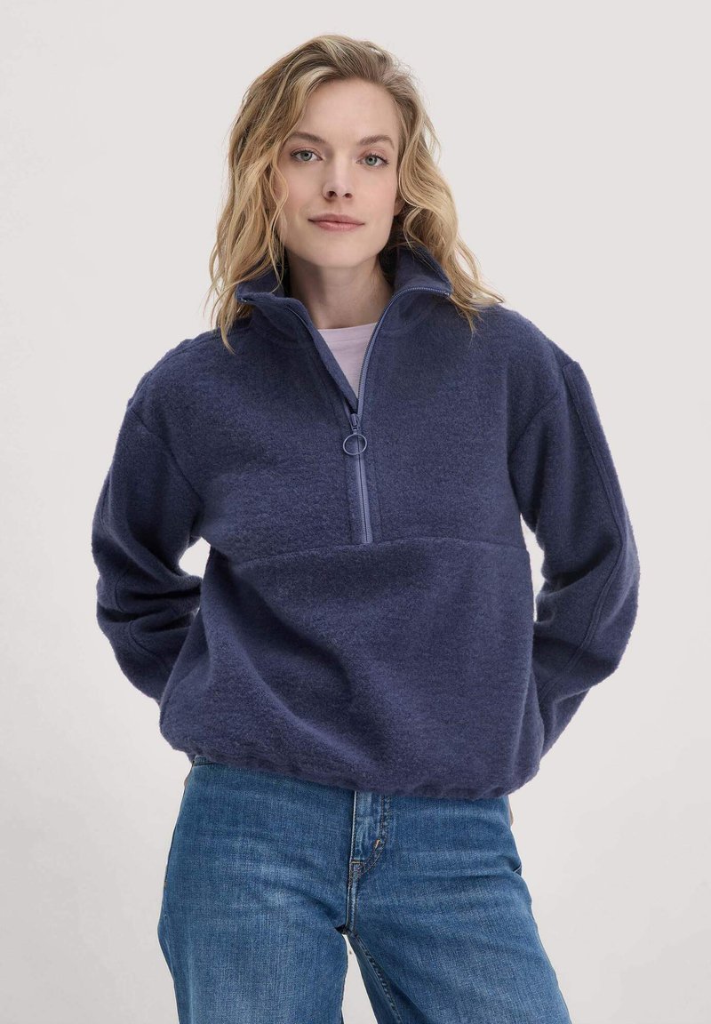 Marineblauer Fleece-Pullover mit einem Viertelreißverschluss, ausgestattet mit einem runden Reißverschluss-Zug, gerippten Bündchen und einem lockeren Schnitt. Kombiniert mit blauen Jeans.