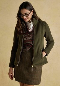 Woman wearing glasses, olive green zip jacket, patterned sweater vest, white ruffled blouse, and brown plaid skirt, smiling and looking down.