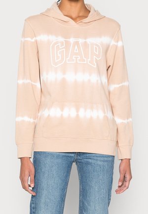 Henkilö pukeutunut beige-valkoiseen liukuvärjättyyn GAP-huppariin, jossa on etutasku, ja sinisiin farkkuihin, seisomassa vaaleaa taustaa vasten.