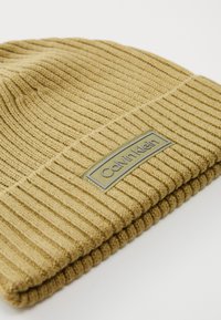 Beige ribbed knit beanie folded at the edge with a rectangular Calvin Klein brand label stitched on the cuff.