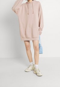 Helles rosa Kapuzensweatkleid mit langen Ärmeln, Fronttasche und Kordelzug. Kombiniert mit weißen Knöchelsocken und cremefarbenen Sneakers.