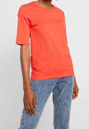 Personne portant un t-shirt orange vif à manches courtes et un jean en denim bleu clair sur un fond uni.