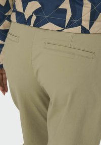 Beige kalhoty s hladkým povrchem, vybavené dvěma zadními kapsami a přiléhavým střihem. Pás je částečně viditelný.