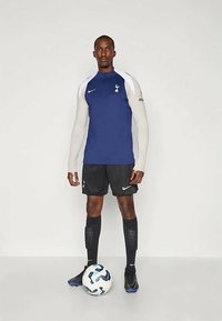 Marineblå og hvid langærmet trøje med høj krave, sorte shorts og sorte sokker. Spilleren holder en hvid fodbold med blå detaljer.