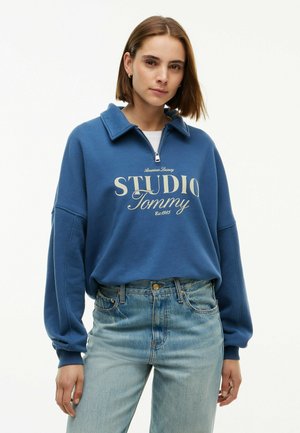 Ung kvinde med kort brunt hår iført en blå lynlås-sweatshirt med teksten "Studio Tommy" og lyseblå jeans, stående mod en hvid baggrund.