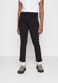 Jeans skinny pretos com cintura média, apresentando uma textura suave e leve elasticidade. Combinados com sapatos loafers verdes-oliva e meias brancas.