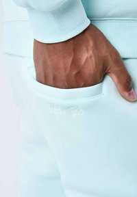 Pantalones de chándal azul claro con un bolsillo lateral. Presenta una cintura acanalada y un logo bordado en blanco. Tejido suave y con textura.