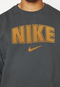 Donkergrijze sweatshirt met grote, vetgedrukte oranje "NIKE" tekst en een klein oranje Nike swoosh logo. Ronde halslijn en zachte textuur.