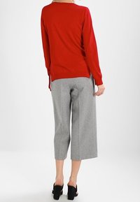 Pull en laine rouge avec un col arrondi et des fentes sur les côtés, associé à un pantalon large gris. Des chaussures à talons carrés noires complètent le look.