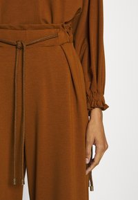 Ensemble en tissu texturé marron avec taille cintrée. Présente des poignets froncés et des cordons tressés avec des embouts métalliques.