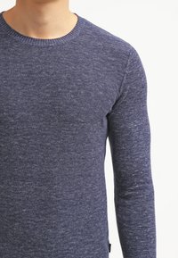 Granatowy dzianinowy sweter z okrągłym dekoltem i długimi rękawami; miękka tkanina z delikatnym melanżowym wzorem i detalem w postaci bocznej metki.