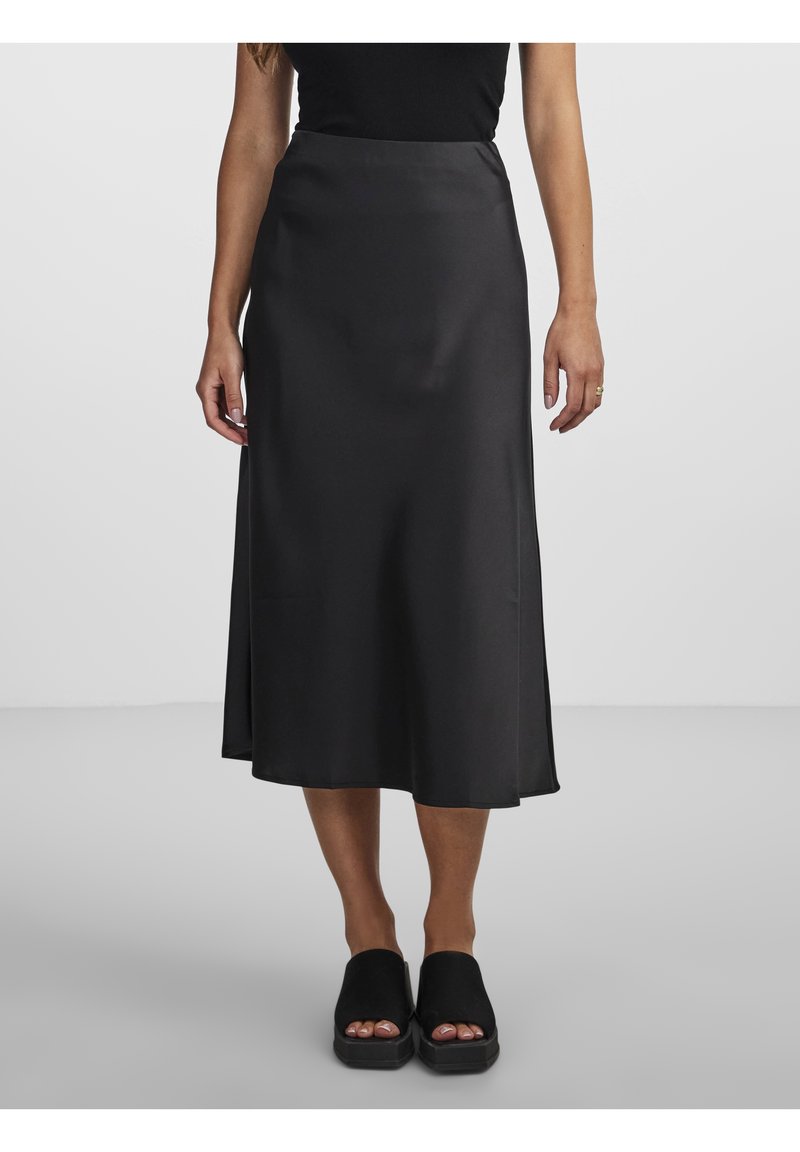 Femme portant une jupe midi en satin noire avec un top noir sans manches et des sandales à plateforme noires à bout ouvert, debout sur un sol gris.
