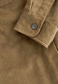 Brown corduroy fabric featuring vertical ridges, with a close-up of a cuff and a engraved metal snap button detail.