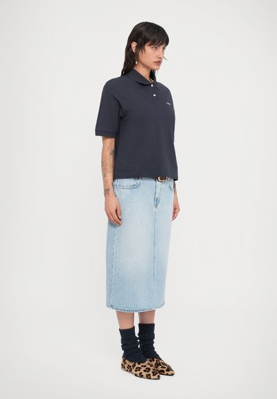 Γυναίκα με μπλε σκούρο πόλο πουκάμισο, ανοιχτό μπλε τζιν μίντι φούστα, μαύρες ζαρτιέρες και μυτερές μπαλαρίνες με λεοπάρ σχέδιο, όρθια πλαγίως.