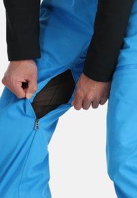 Wasserabweisende blaue Hose mit Reißverschlussöffnung, ausgestattet mit schwarzem Mesh-Futter und einem schlanken Design. Glatte Textur und taillierte Form.