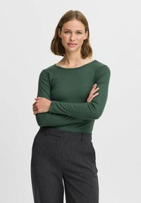 Grünes, geripptes Langarmshirt mit rundem Ausschnitt, kombiniert mit dunkelgrauen, maßgeschneiderten Hosen. Glatte Textur, figurbetontes Design.