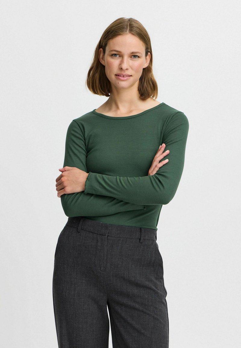 Grünes, geripptes Langarmshirt mit rundem Ausschnitt, kombiniert mit dunkelgrauen, maßgeschneiderten Hosen. Glatte Textur, figurbetontes Design.