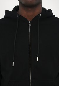Felpa nera con zip realizzata in cotone, dotata di cappuccio con cordino, zipper argento a contrasto e tasche anteriori. Texture liscia.