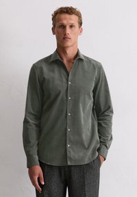 Chemise boutonnée en corduroy vert, avec un col classique, des manches longues et des boutons en ton argent, associée à un pantalon gris texturé.