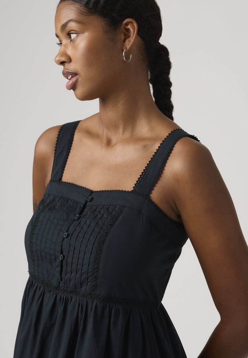 Jeune femme aux cheveux tressés portant des boucles d'oreilles créoles et une robe noire sans manches avec un corsage plissé à boutons.