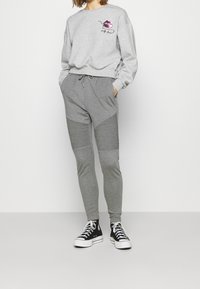 Sudadera gris claro con gráfico de labios impresos y texto, combinada con joggers gris claro que presentan detalles de paneles acanalados. Usada con zapatillas negras.