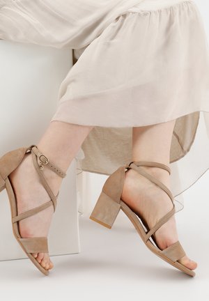 Sandali con tacco in suede beige con tacco a blocco. Presentano cinturini incrociati e un cinturino alla caviglia con un piccolo dettaglio di fibbia dorata.