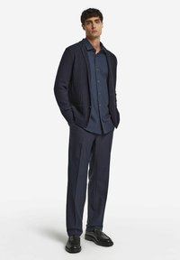 Marine strikket cardigan over en mørkeblå skjorte med knapper, parret med mørke bukser og sorte loafers. Sættet har et rent, struktureret design.
