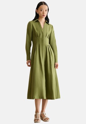 Frau trägt ein olivgrünes Midikleid mit langen Ärmeln, einem Kragen und plissierter Taille, kombiniert mit beigen Keilsandalen.