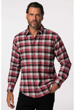 Mand med skæg iført rød, hvid og sort ternet skjorte med knapper og sorte bukser, stående med den ene hånd i lommen mod en hvid baggrund.