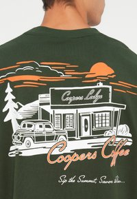 Camiseta de color verde oscuro con un diseño gráfico de una cabaña de café con montañas y un sol, utilizando contornos en blanco y naranja.