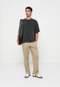 Young man wearing black oversized T-shirt, beige pants, white sneakers with black stripes, holding black shoulder bag against white background.