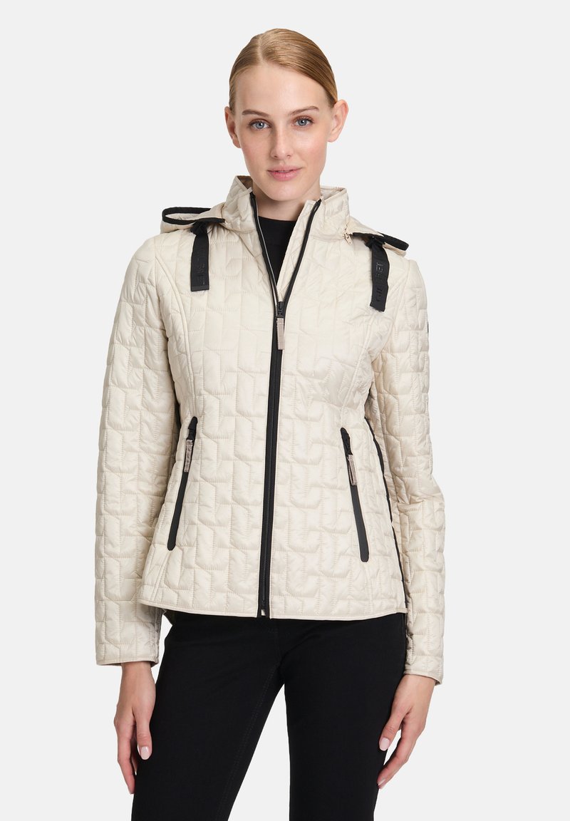 Donna che indossa una giacca trapuntata beige con zip nera e dettagli neri, in piedi contro uno sfondo bianco semplice.