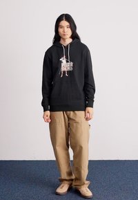 Black hoodie with a front graphic, drawstring hood, kangaroo pocket; paired with beige cargo pants and light brown shoes.