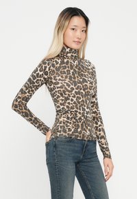 Sieviete ar gariem blondiem matiem, ģērbusies leopardu raksta augstākajā kaklā un zilos džinsos, pozējot ar vienu roku kabatā baltā fona priekšā.
