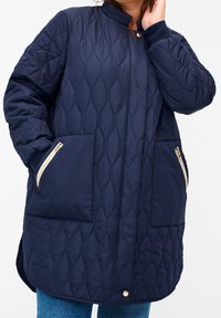 Marineblå, quilted lang jakke med ståkrave, to lynlåslommer i siden med cremefarvede detaljer og en afslappet pasform.