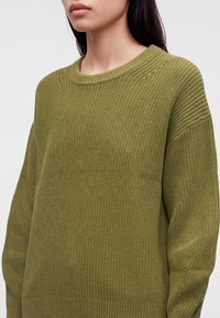 Femme portant un pull en tricot texturé vert olive avec un col rond, visible des épaules jusqu'au milieu du torse sur un fond blanc.