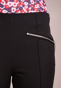 Pantaloni neri con una tasca laterale con zip e hardware argentato, realizzati in un tessuto liscio e strutturato. Top a fantasia floreale visibile sopra.