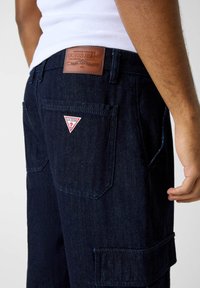 Ciemne dżinsowe spodnie typu cargo z naszywką z logo Guess z tyłu, widocznym przeszyciem i głębokimi kieszeniami. Tkanina ma teksturowany, wytrzymały wygląd.