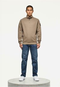Lichtbruine fleece pullover met een hoge kraag en een halve ritssluiting, gedragen met blauwe denim jeans en witte sneakers, op een ronde platform.