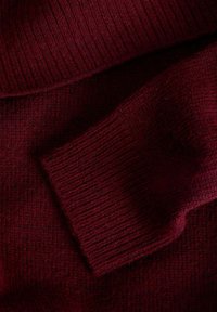 Vīna sarkanā krāsā adīts džemperis ar ribotu tekstūru, salocītu apkakli un izteiktu aproces detaļu. Materiāls šķiet mīksts un silts.