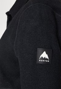Giacca in pile nera con chiusura zip, tessuto testurizzato e un'etichetta rettangolare con un logo di montagna e il testo "Burton."