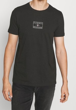 Homme portant un t-shirt noir Tommy Hilfiger avec un patch logo sur la poitrine, associé à un jean noir, debout devant un fond clair uni.