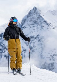 Ski-uitrusting met een zwart-orenjack, bruine broek en skistokken. Bevroren landschap met besneeuwde bergen op de achtergrond.